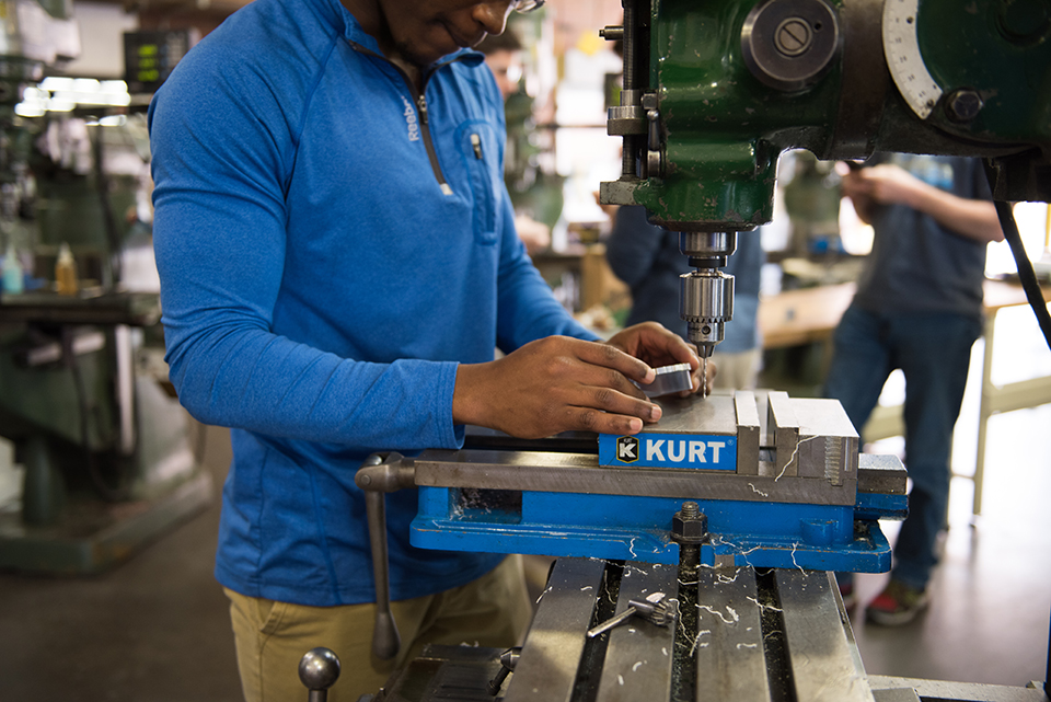 Student wearing a blue shirt working with a drill press.